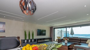 A modern living room with a large window, a dining table with fruit, a sofa, and a TV mounted on the wall. - Lorena Stylish Sea View Villa (Crikvenica)