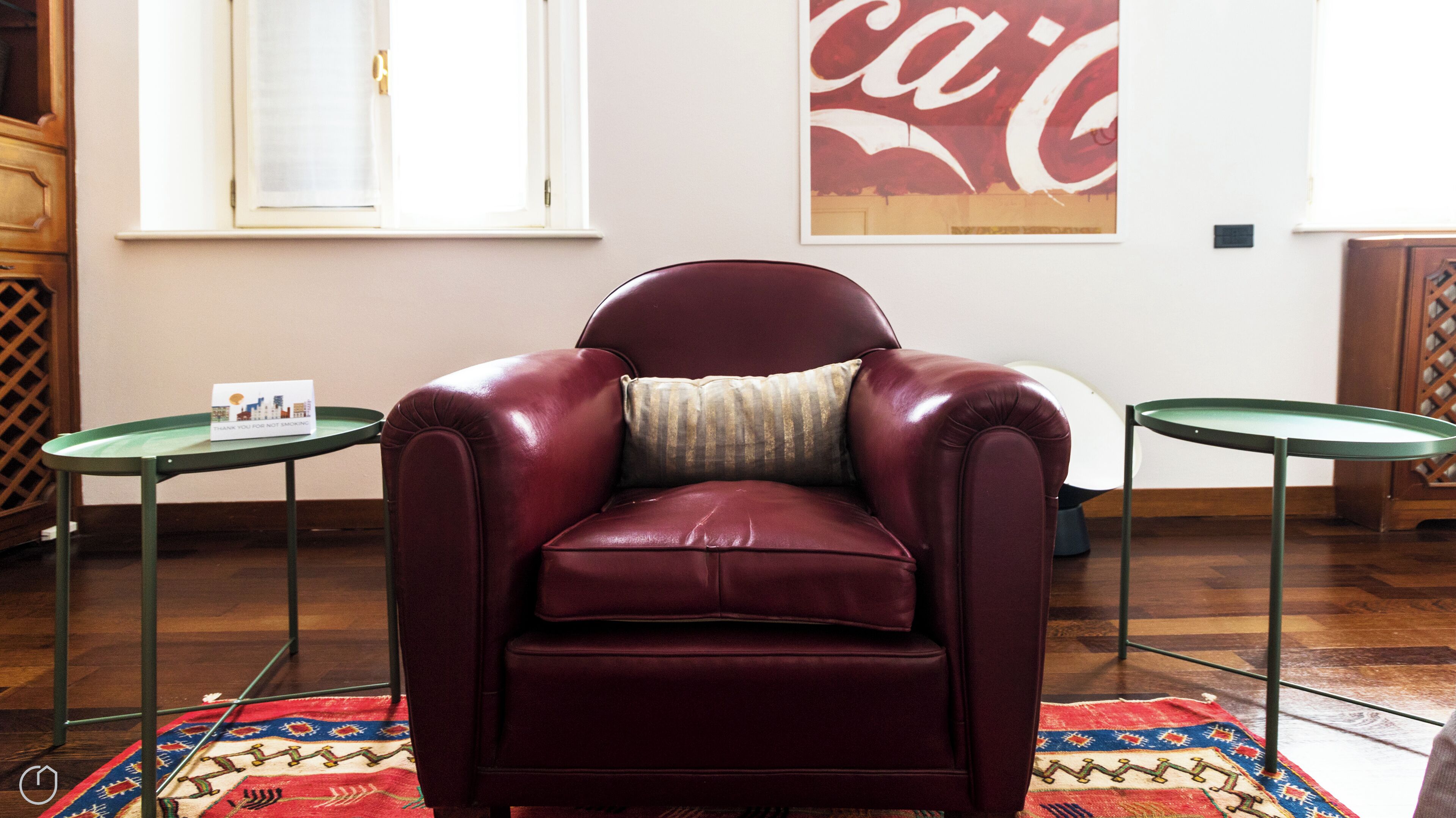 A maroon leather armchair with a striped pillow, positioned on a patterned rug. There are two small tables with green tops, one on each side of the chair. A large Coca-Cola poster is visible on the wall behind the chair.
