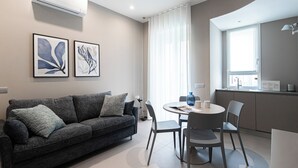 A modern living room with a dark grey sofa, a dining table with chairs, and a kitchen area with a window. - Italianway Comfort - Maderno 4 (Milano)
