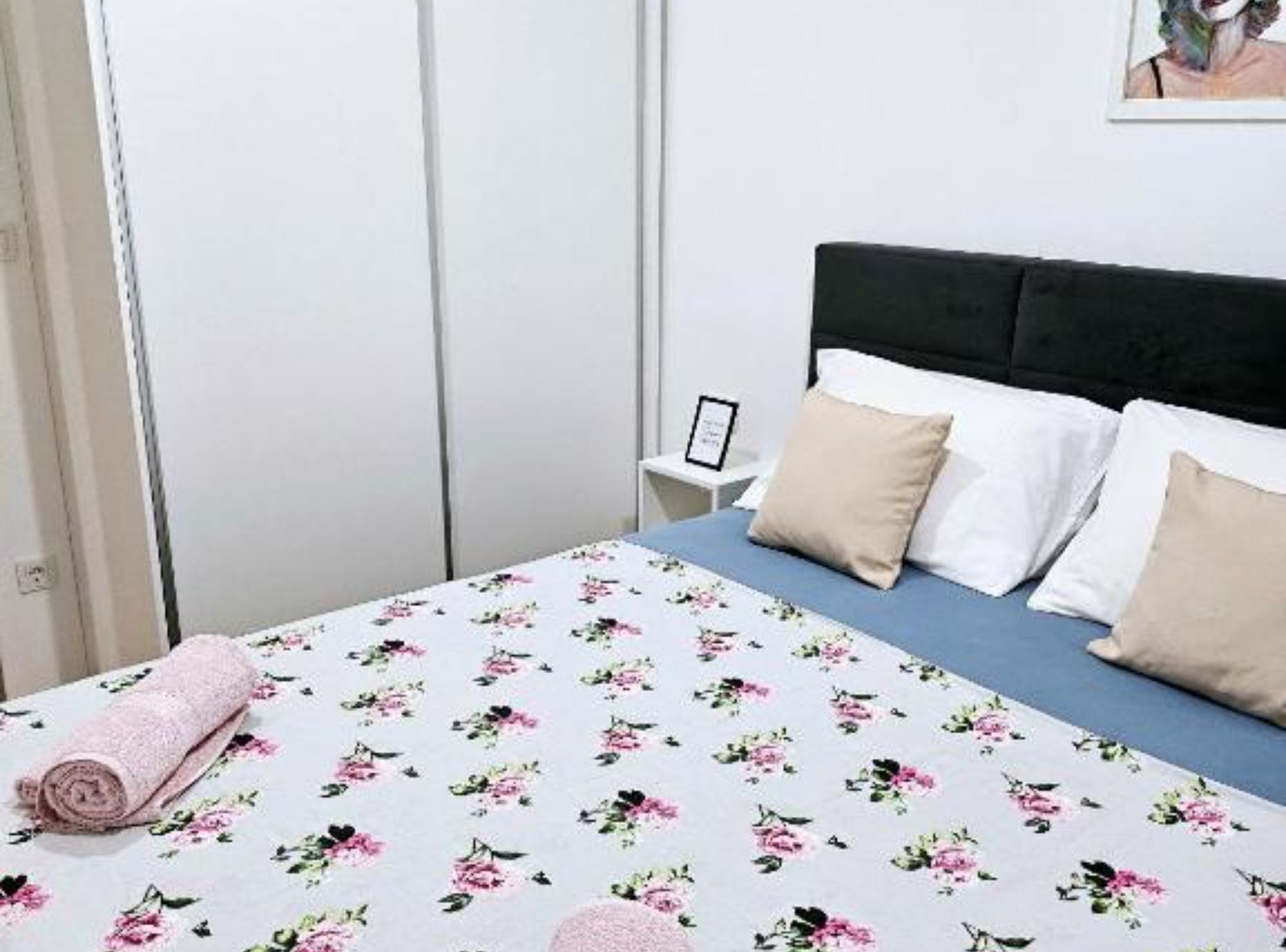 Una cama bien hecha con una colcha floral, una toalla enrollada y un cuadro en la mesita de noche.