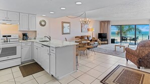A modern kitchen with white cabinetry, a built-in oven, and a coffee maker. There is a dining area with a table and chairs, and a living room with a sofa and a TV. The room has a large window with a view of the ocean. - Gulf Front Getaway at Plumlee Vacation Rental Chateaux 107 (Indian Shores)