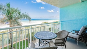A balcony with a view of the beach, a round table, wicker chairs, and a blue wall. - Gulf Front Getaway at Plumlee Vacation Rental Chateaux 107 (Indian Shores)