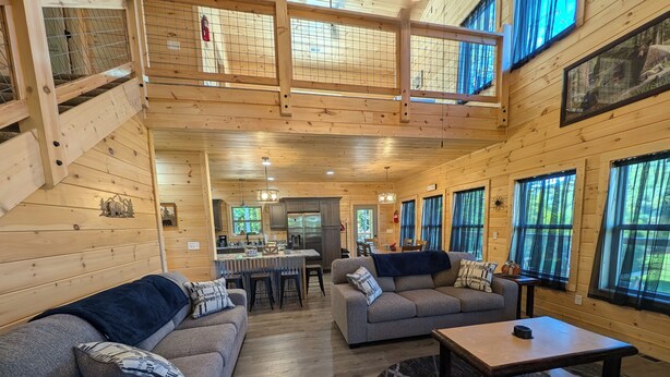 A spacious cabin with a high ceiling, wooden walls, and large windows. It features a living area with a sofa, a coffee table, and a dining area with a kitchen in the background. - 3 Master Suites, Easy Access w/View, Game&Theater Rms, Fire Pit, & Pool Access (Sevierville)
