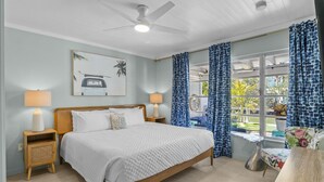 Une chambre à coucher comprenant un lit, deux tables de chevet, un ventilateur de plafond, une fenêtre avec des rideaux et une vue sur l’extérieur.