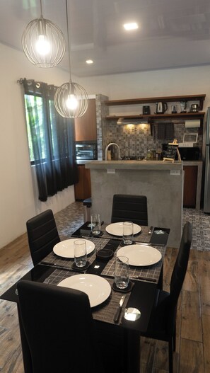 Un coin repas avec une table dressée pour quatre personnes, une cuisine en arrière-plan et une vue sur l’extérieur par une fenêtre.