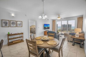 A modern living room with a dining area, large windows with a view, and a ceiling fan. - Ariel Dunes II 907 *Beach views *Pools* (Miramar Beach)