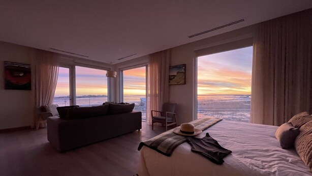 Een ruime slaapkamer met een groot bed, een bank en uitzicht op de ondergaande zon door grote ramen.