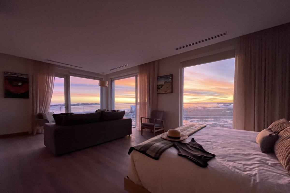 Ein geräumiges Schlafzimmer mit einem großen Bett, einer Couch und Blick auf den Sonnenuntergang durch große Fenster.
