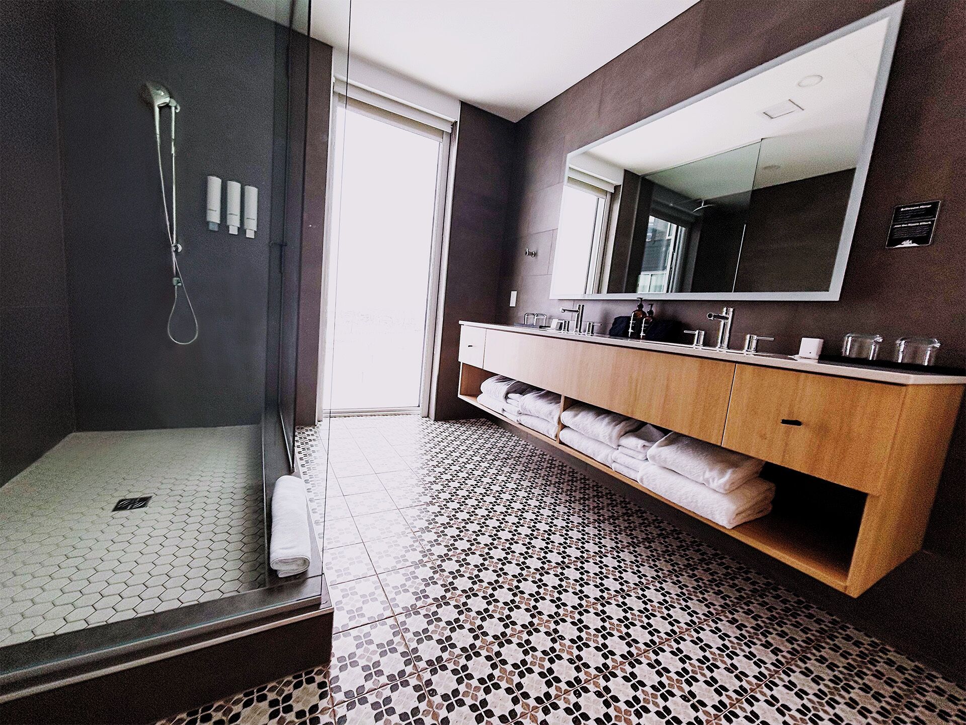 A modern bathroom with a glass-enclosed shower, a wooden vanity with a large mirror, and a patterned floor.