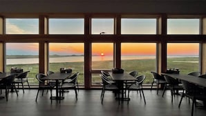 Een eetruimte met meerdere tafels en stoelen, grote ramen met uitzicht op een ondergaande zon boven het water.