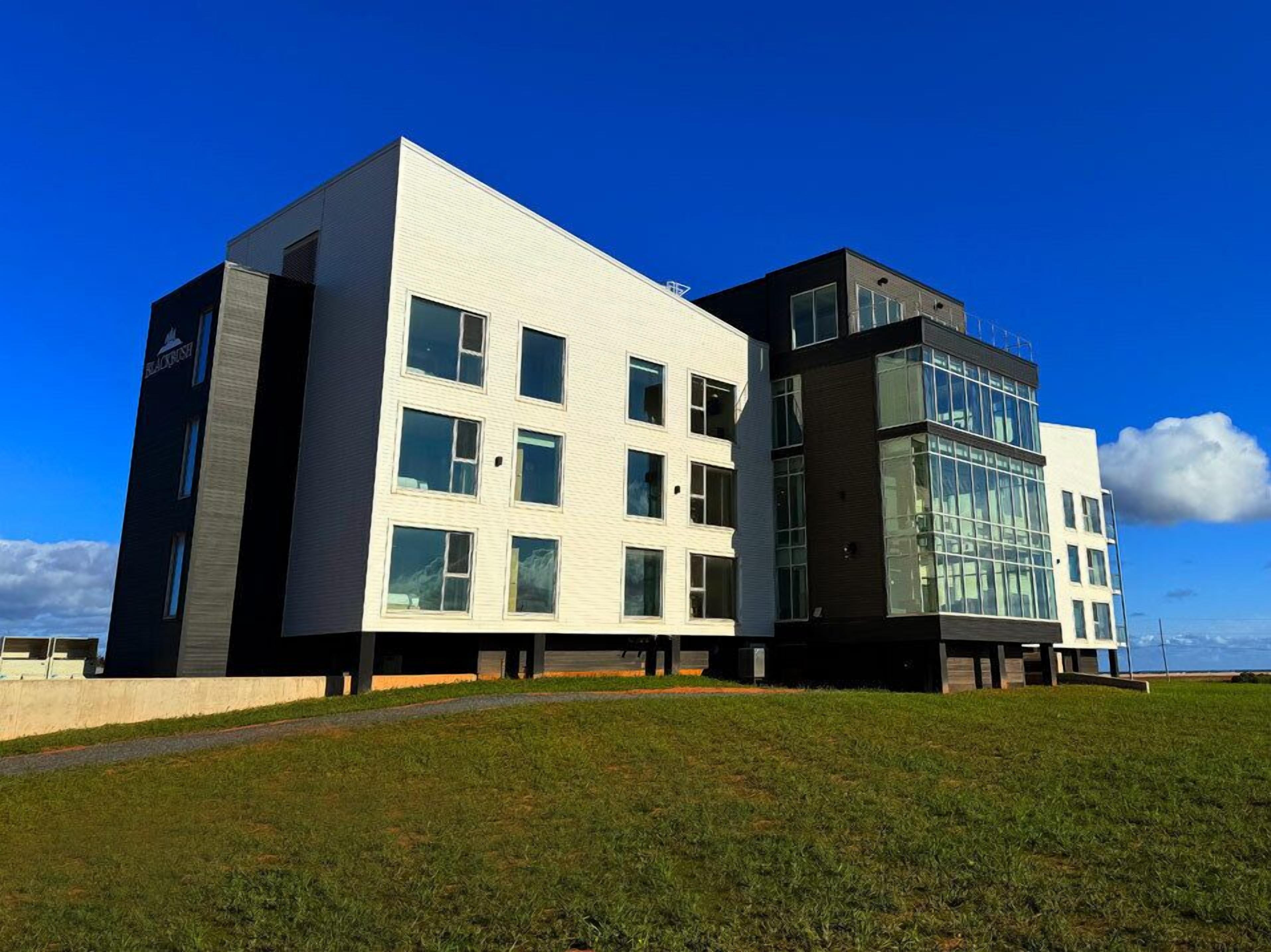 A modern building with a white facade and large windows, located on a grassy hill with a clear blue sky.
