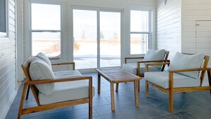 Een zonnige kamer met een grote schuifglazen deur die uitkijkt op een besneeerd landschap. Binnen staan twee houten stoelen met kussens en een klein houten tafeltje.