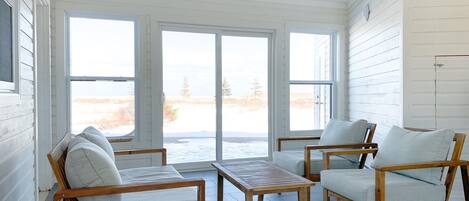 A sunlit room with a large sliding glass door leading to a view of a snowy landscape. Inside, there are two wooden chairs with cushions and a small wooden table.
