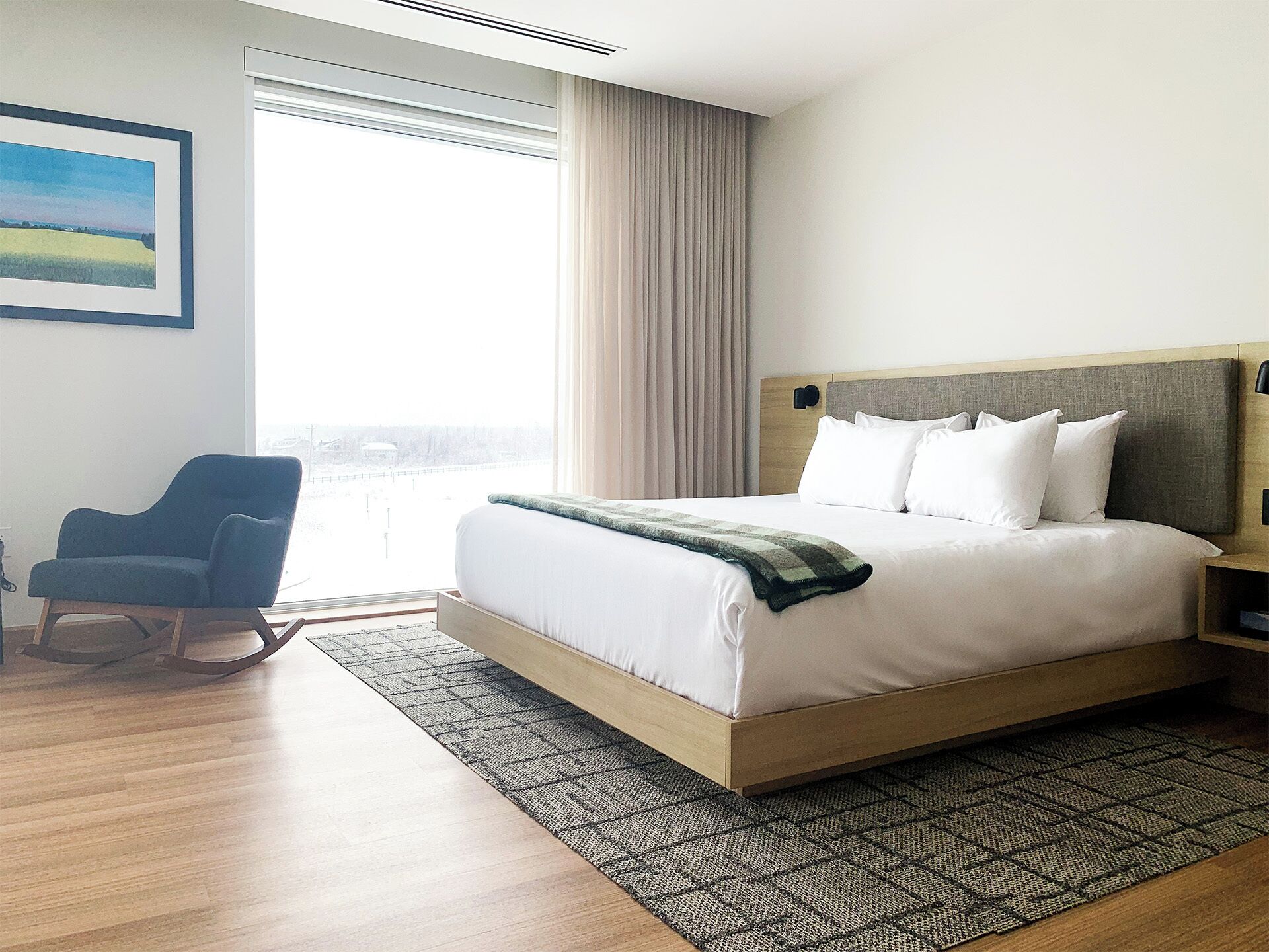 Ein Hotelzimmer mit einem großen Bett, einem Sessel, Blick auf die Stadt und einem Gemälde an der Wand.