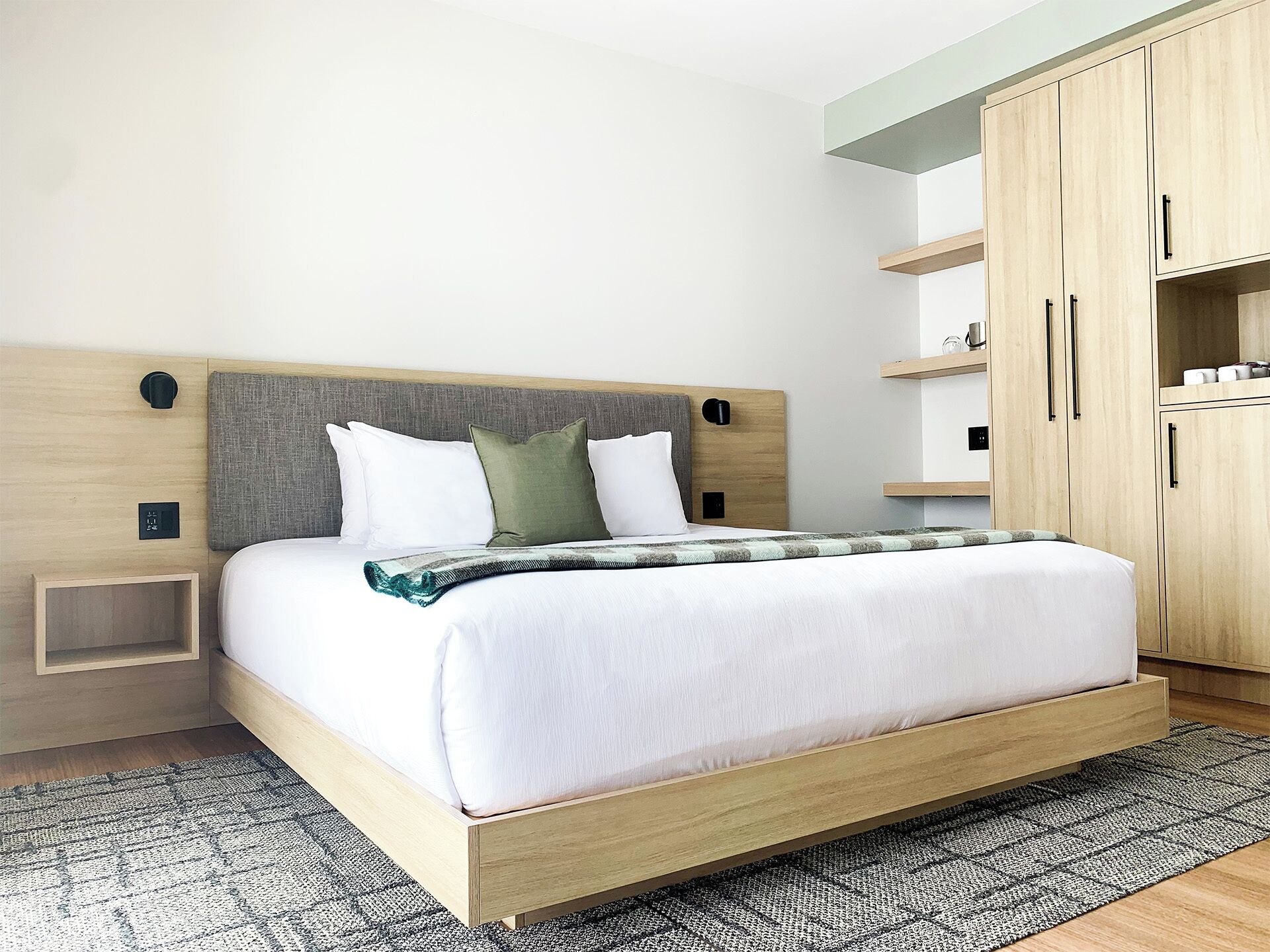 A modern hotel room with a wooden bed, a grey headboard, and built-in wooden shelving with a small bedside table.