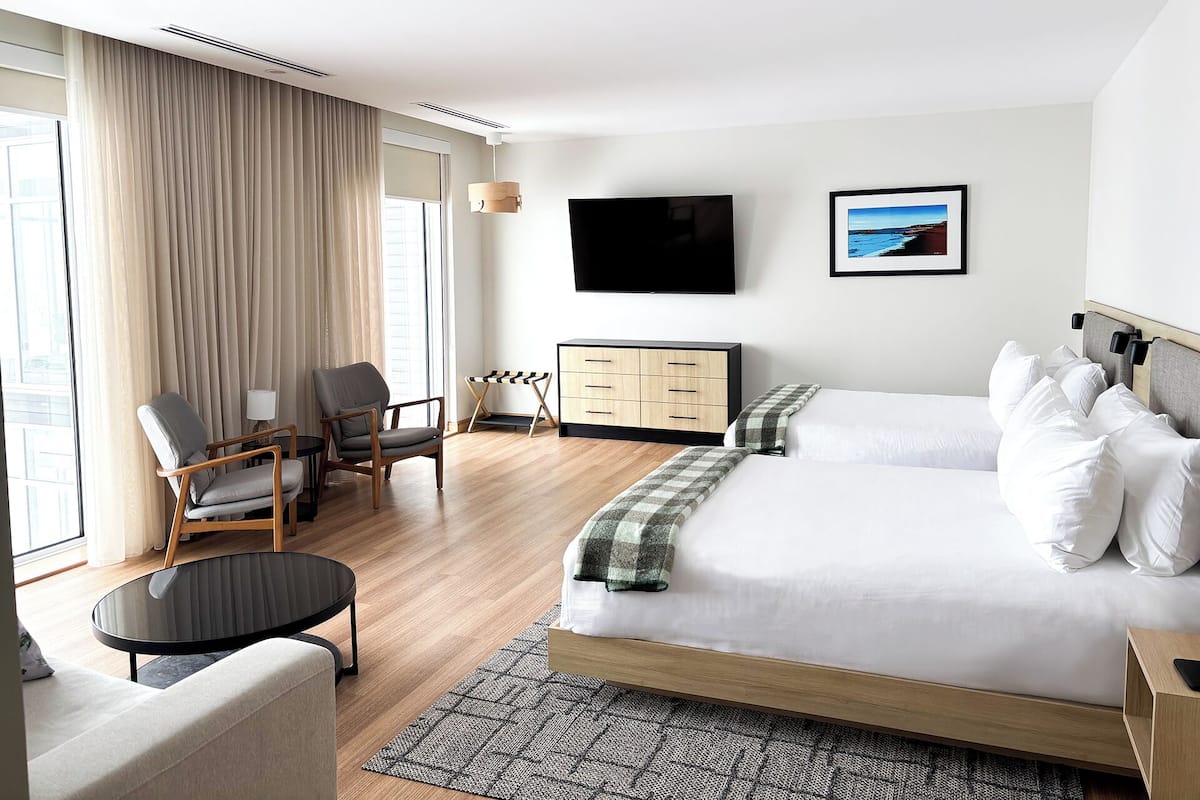Ein modernes Hotelzimmer mit einem großen Bett, zwei Sesseln, einem kleinen Tisch, einem Fernseher und einem Holzkasten.