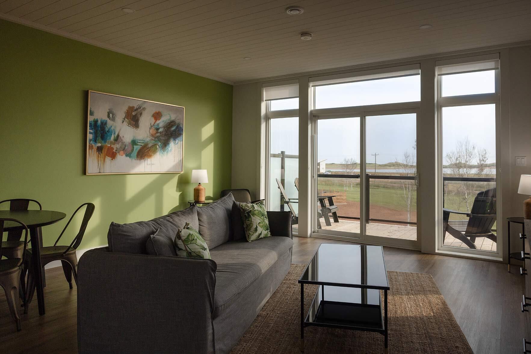 A spacious living room with a grey sofa, a coffee table, and a painting on the wall. There is a large sliding glass door leading to an outdoor area with a barbecue grill.