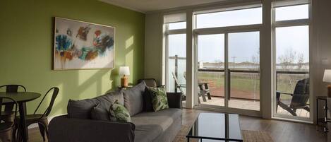 A spacious living room with a grey sofa, a coffee table, and a painting on the wall. There is a large sliding glass door leading to an outdoor area with a barbecue grill.