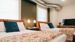 A hotel room with two beds, each with a floral bedspread, a nightstand with a lamp, a television on a wooden stand, and a large window with blinds. - Westphalia Inn (Westphalia)