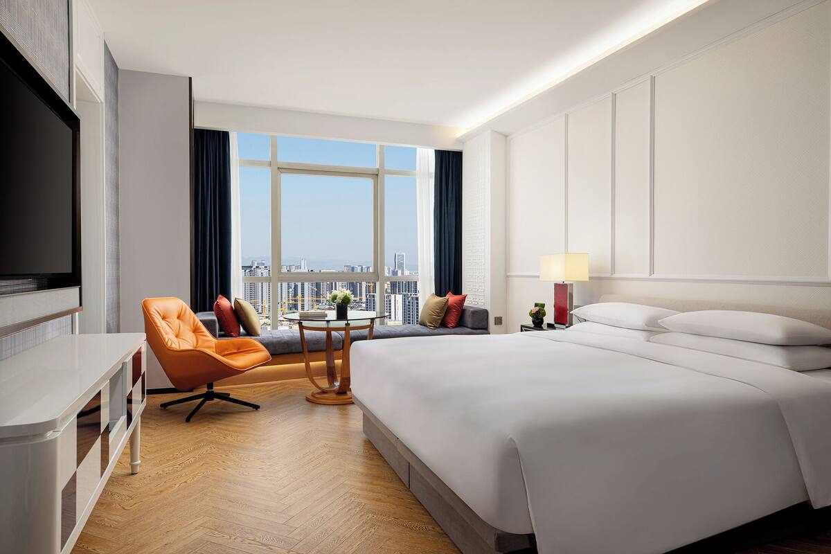 Ein Hotelzimmer mit einem großen Bett, einer Sitzecke mit Sessel und Tisch, einem Fernseher und Blick auf die Stadt.