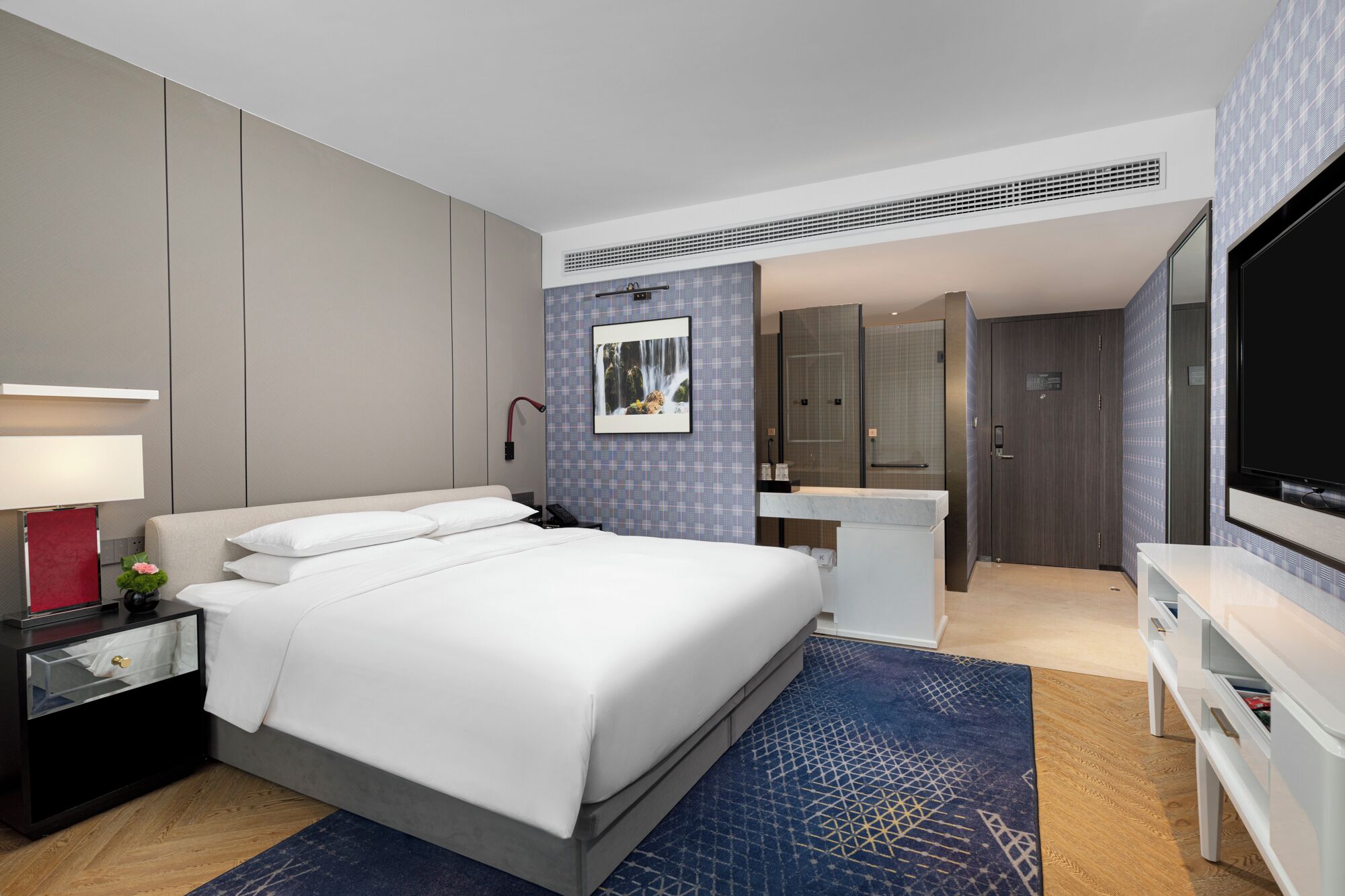 Quarto de hotel moderno com uma cama grande, mesa de cabeceira com abajur, televisão, banheiro com chuveiro e uma escrivaninha.