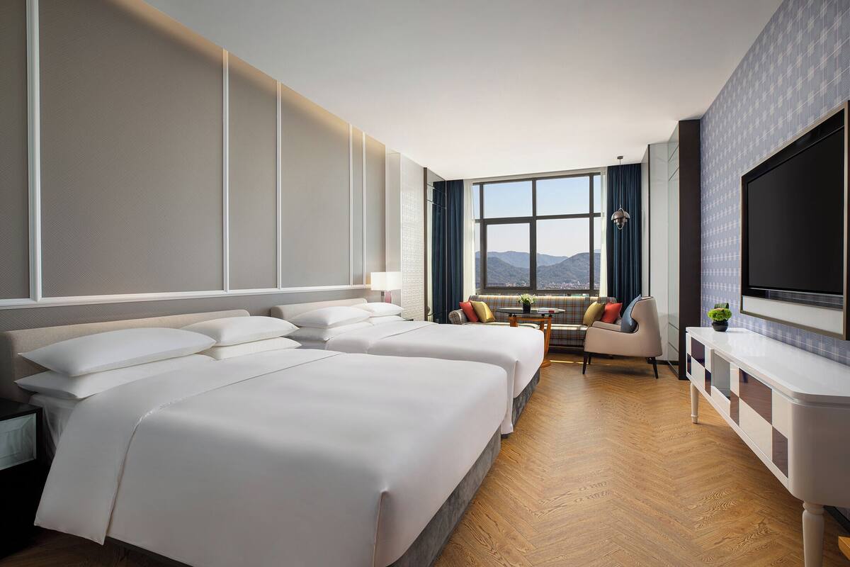 Ein Hotelzimmer mit zwei Betten, einer Sitzecke mit Blick und einem an der Wand befestigten Fernseher.
