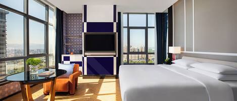 A hotel room with a large bed, a seating area with a table and chairs, a TV, and a city view through large windows.