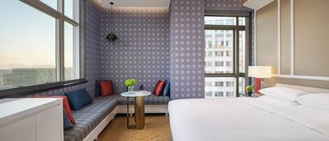 A hotel room with a large bed, a seating area with a view, and a small table with a lamp.