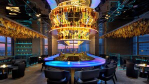 Eine moderne Bar mit einer zentralen Spirituosenpräsentation, bequemen Sitzgelegenheiten und großen Fenstern mit Blick auf die Stadt.