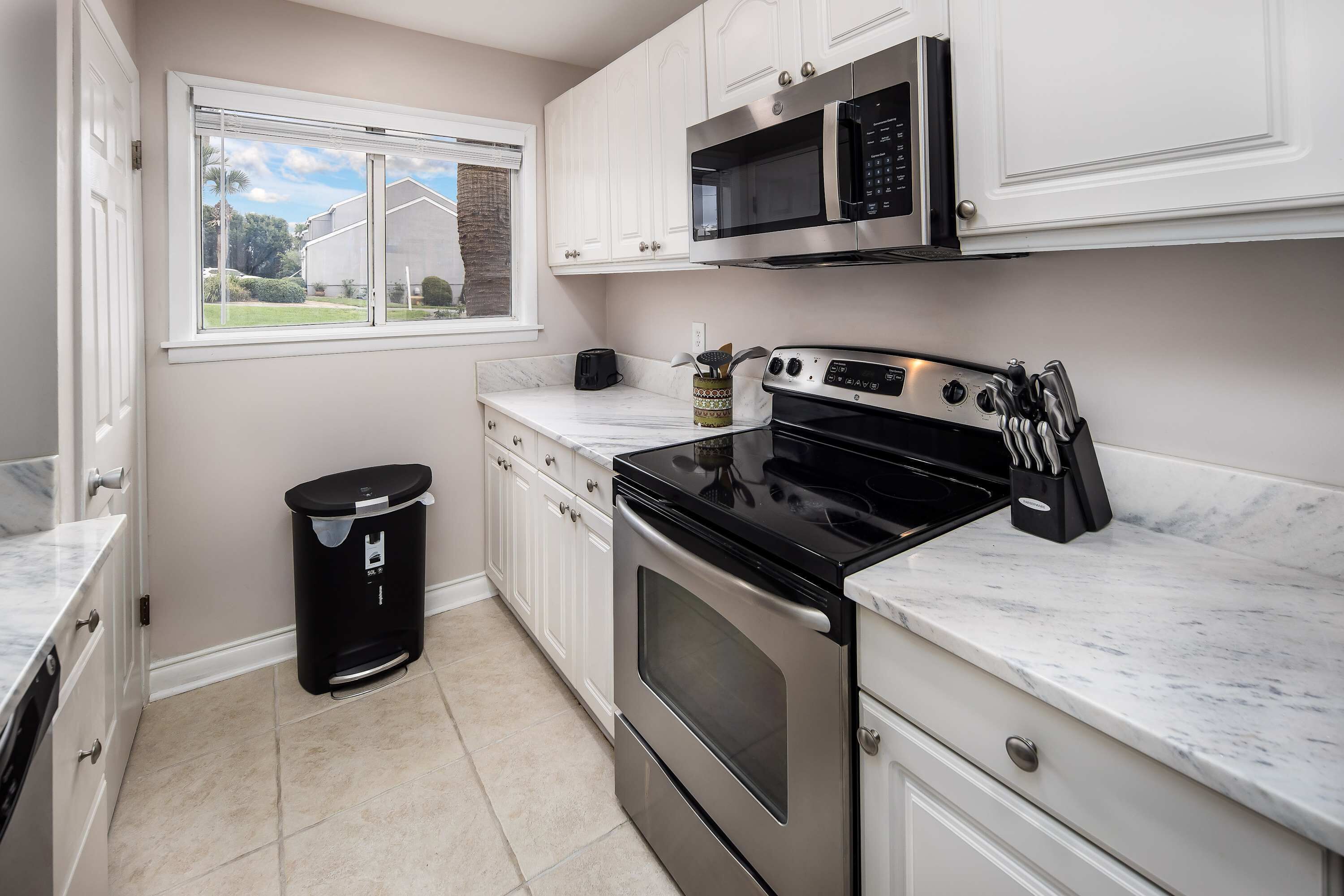 A kitchen with white cabinets, a stainless steel oven, a microwave, and a window with a view of a house and trees.