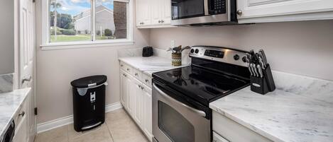 A kitchen with white cabinets, a stainless steel oven, a microwave, and a window with a view of a house and trees.