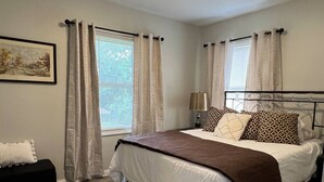 Un dormitorio con cama, cortinas, una lámpara de noche, una mesita y un cuadro en la pared.