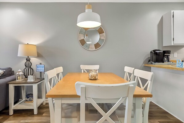 A dining area with a wooden table and white chairs, a hanging light fixture, a coffee maker, and a small side table with a lamp and decorative items.