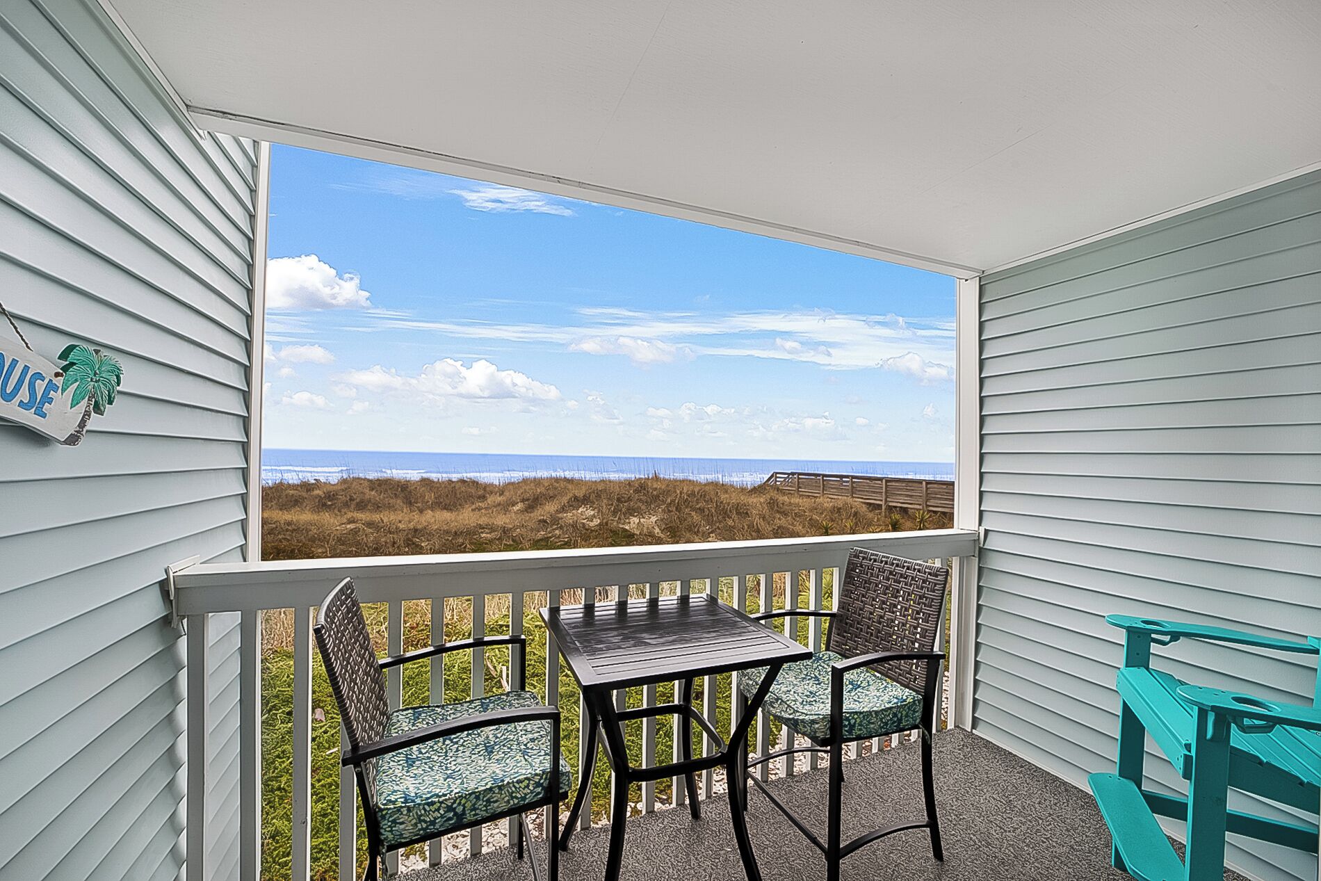 A balcony with a view of the beach, a table, two chairs, and a sign on the wall.