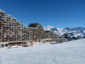 Un hotel de varios pisos con balcones, una pista de esquí en primer plano y montañas nevadas al fondo.