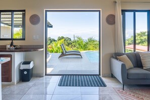 A modern living room with a sliding glass door leading to a pool area, a sofa, a small kitchen area, and a trash bin. - Casa Atardecer (Sámara)