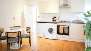 A compact kitchen with a washing machine, oven, and a dining area with chairs and a table. - Fun Energetic Oasis in Islington - Sleeps6 (London)