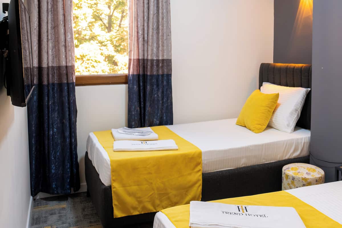 A hotel room with two beds, a window with curtains, an air conditioning unit, a wall-mounted lamp, and a small round table with a floral tablecloth.