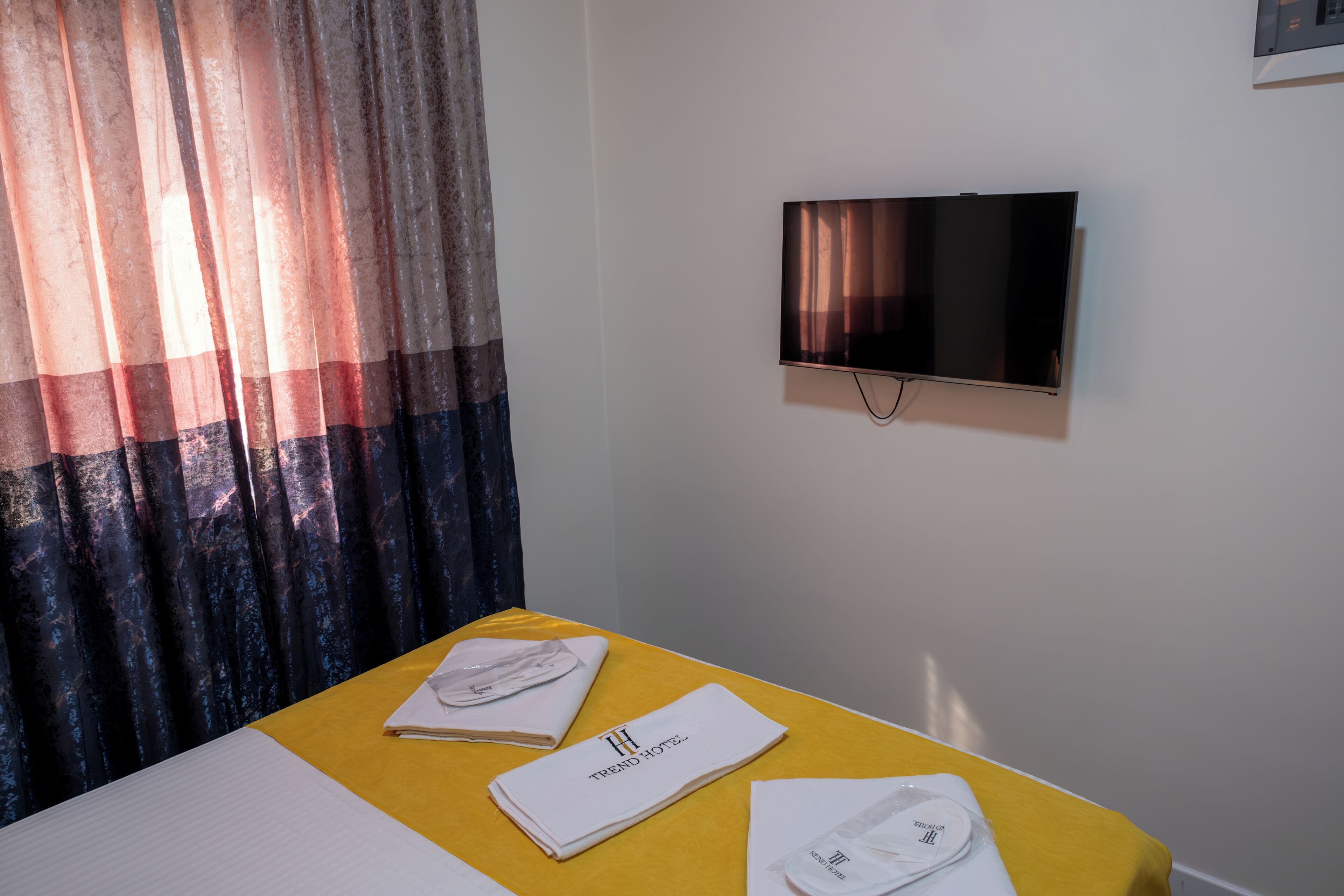 A hotel room with a bed, a TV mounted on the wall, and a table with a folded towel and a white cloth.