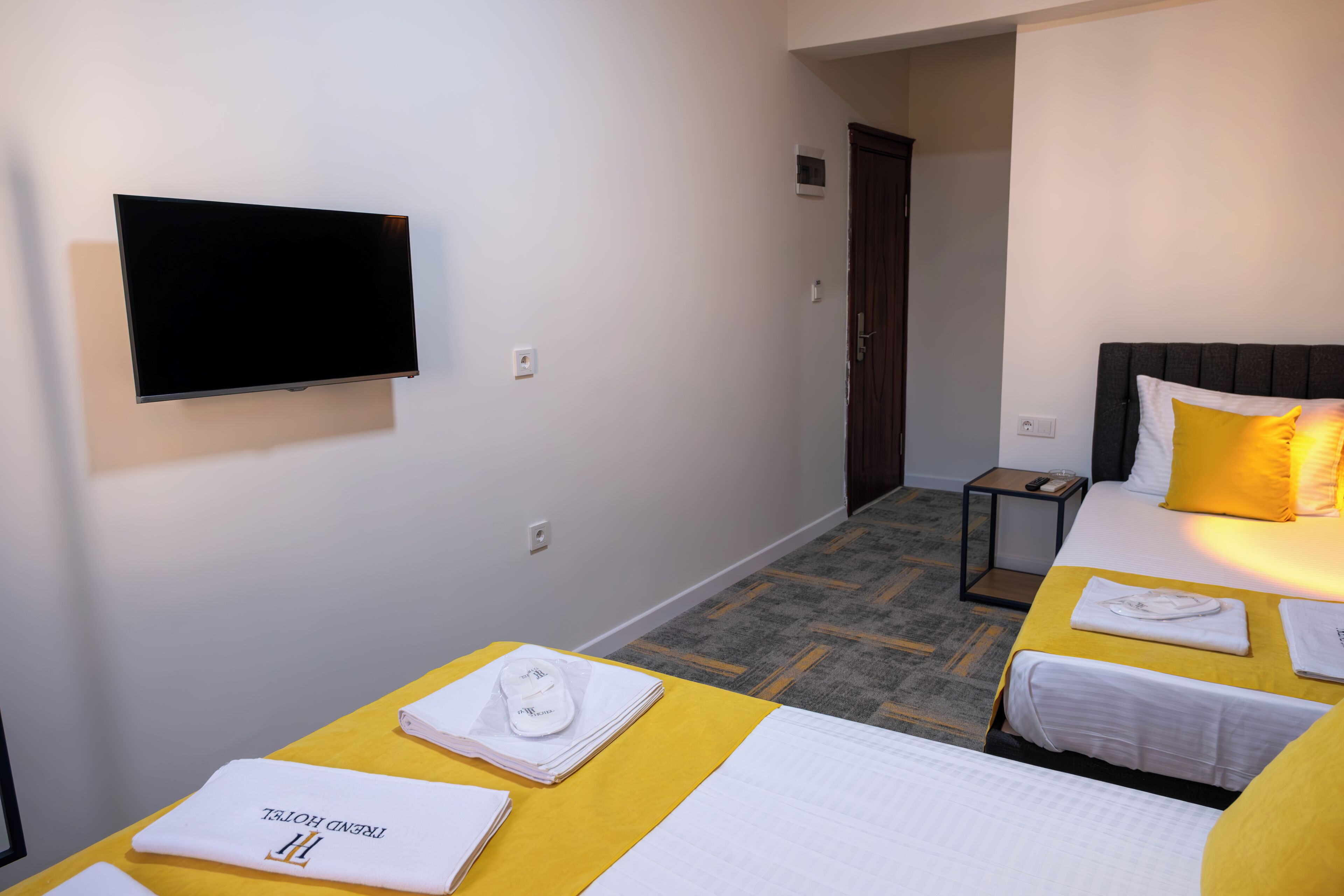 Habitación de hotel con dos camas, un televisor montado en la pared y una mesita de noche pequeña con un control remoto.