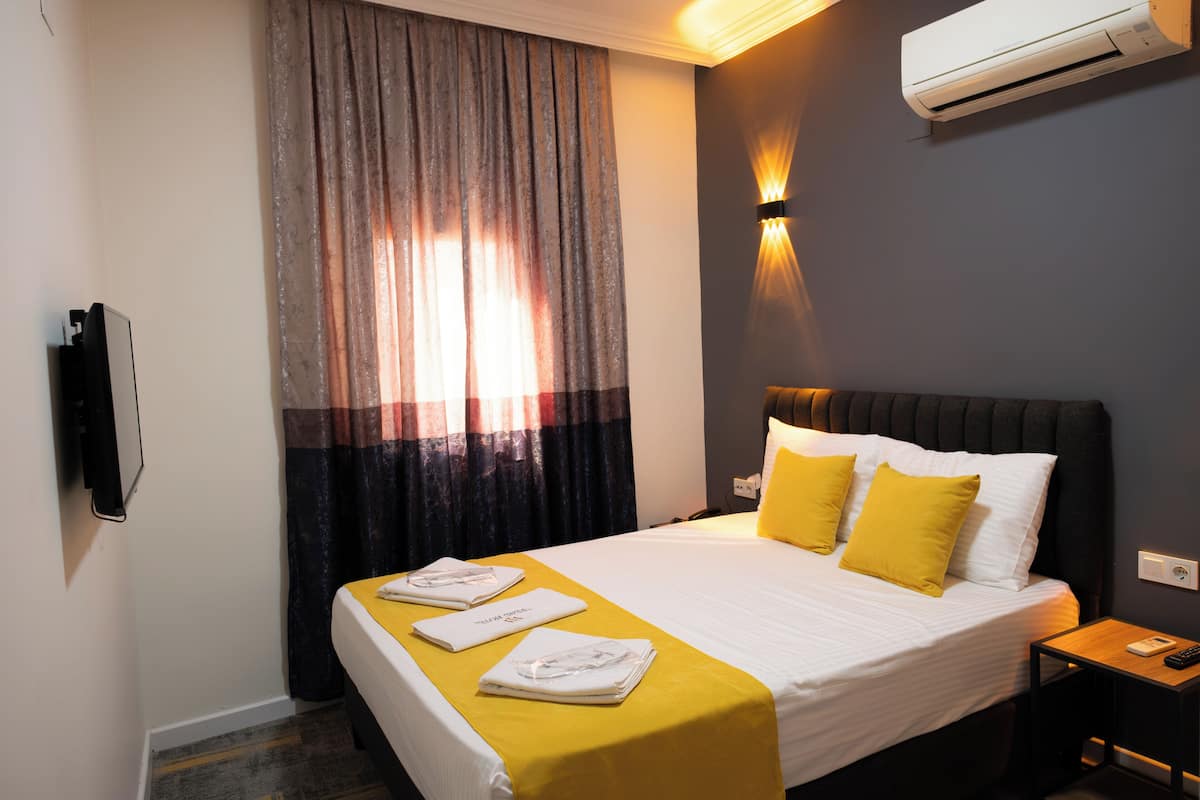 A hotel room with a bed, a bedside table with a lamp, an air conditioner, and a TV on the wall.