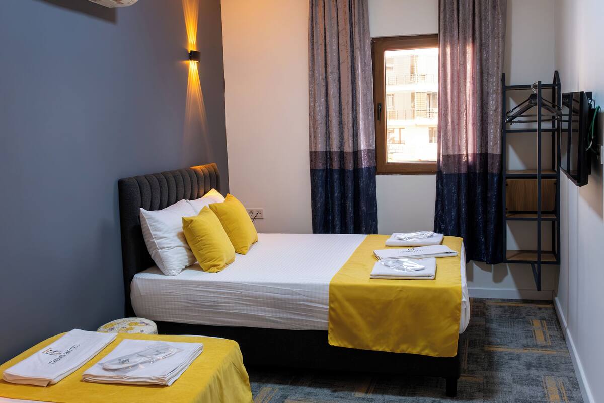 A hotel room with two beds, a window with curtains, an air conditioning unit, a ceiling light, and a wall-mounted TV.