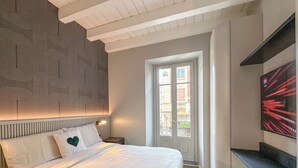 Une chambre à coucher comprenant un lit, une table de chevet, un téléviseur fixé au mur et une fenêtre donnant sur des immeubles.