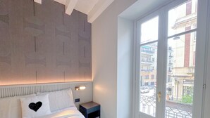 Une chambre à coucher comprenant un lit, une table de chevet, un mur orné d’un motif géométrique et une fenêtre donnant sur des immeubles.