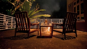 Two wooden chairs and a small table with a fire between them, set on a balcony with a railing and some foliage in the background. - Kilimanjaro Foothills Getaway (Marangu)