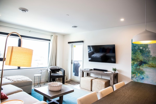 A modern living room with a sofa, ottoman, coffee table, and a dining table. There is a TV mounted on the wall and a painting on the right side.