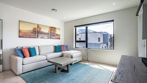 Una sala de estar moderna con un sofá, una mesa de centro y un ventanal con vistas a un edificio y montañas.