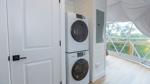 A laundry room with stacked washers and dryers, white walls, and a large window with a view of greenery. - Stunning view! Hot Tub, Privacy & Relaxation (Raywick)