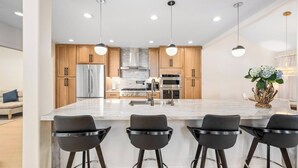 Cocina moderna con una isla central, taburetes de bar, armarios de madera, electrodomésticos de acero inoxidable e iluminación colgante.