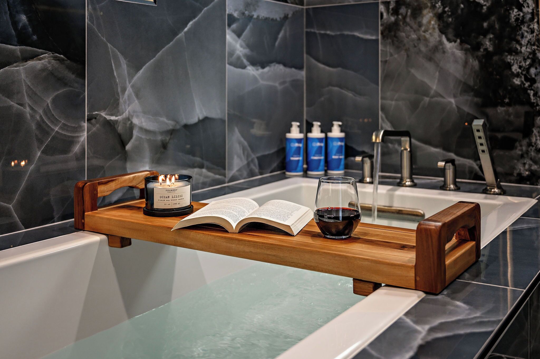 A modern bathroom with a marble wall, a wooden tray holding a book, a candle, and a glass of red wine.
